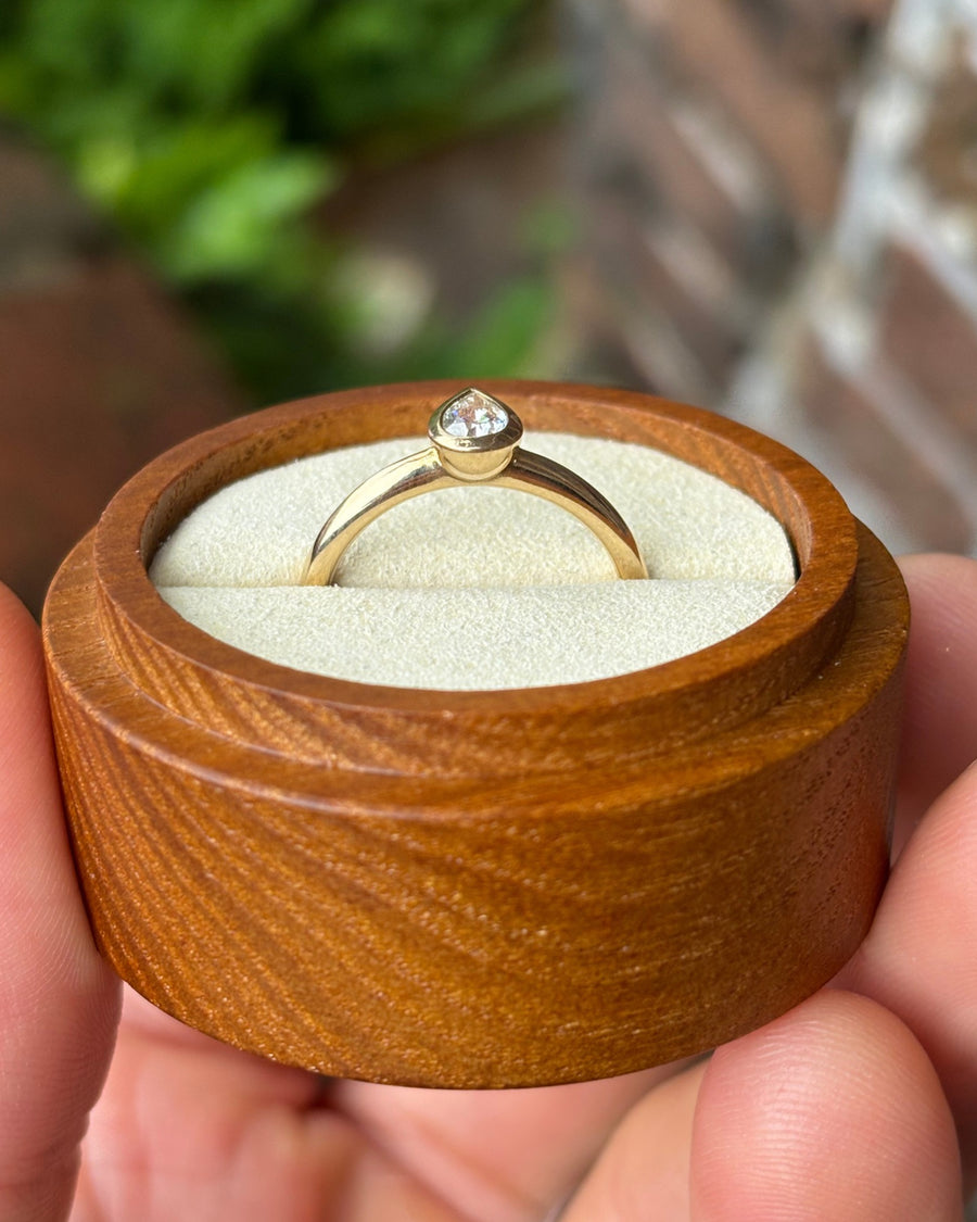 Pear cut diamond engagement ring in a gold bezel setting inside a round wooden box.