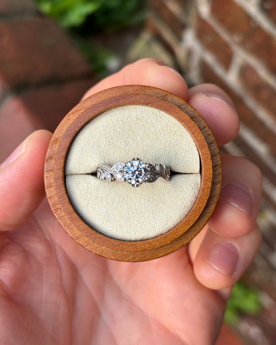 Round diamond engagement ring in a silver-toned metal band with vintage leaf-inspired engravings in a wooden box.
