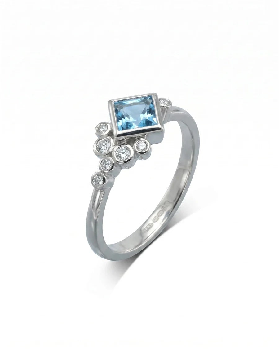 Square blue gemstone engagement ring in white metal with a cluster of small round diamonds.