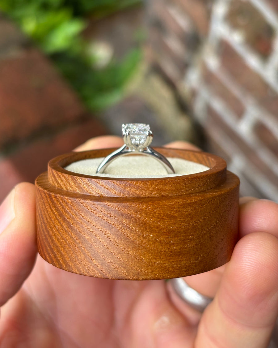Emerald cut diamond engagement ring with a hidden halo in a silver band held in a wooden box.