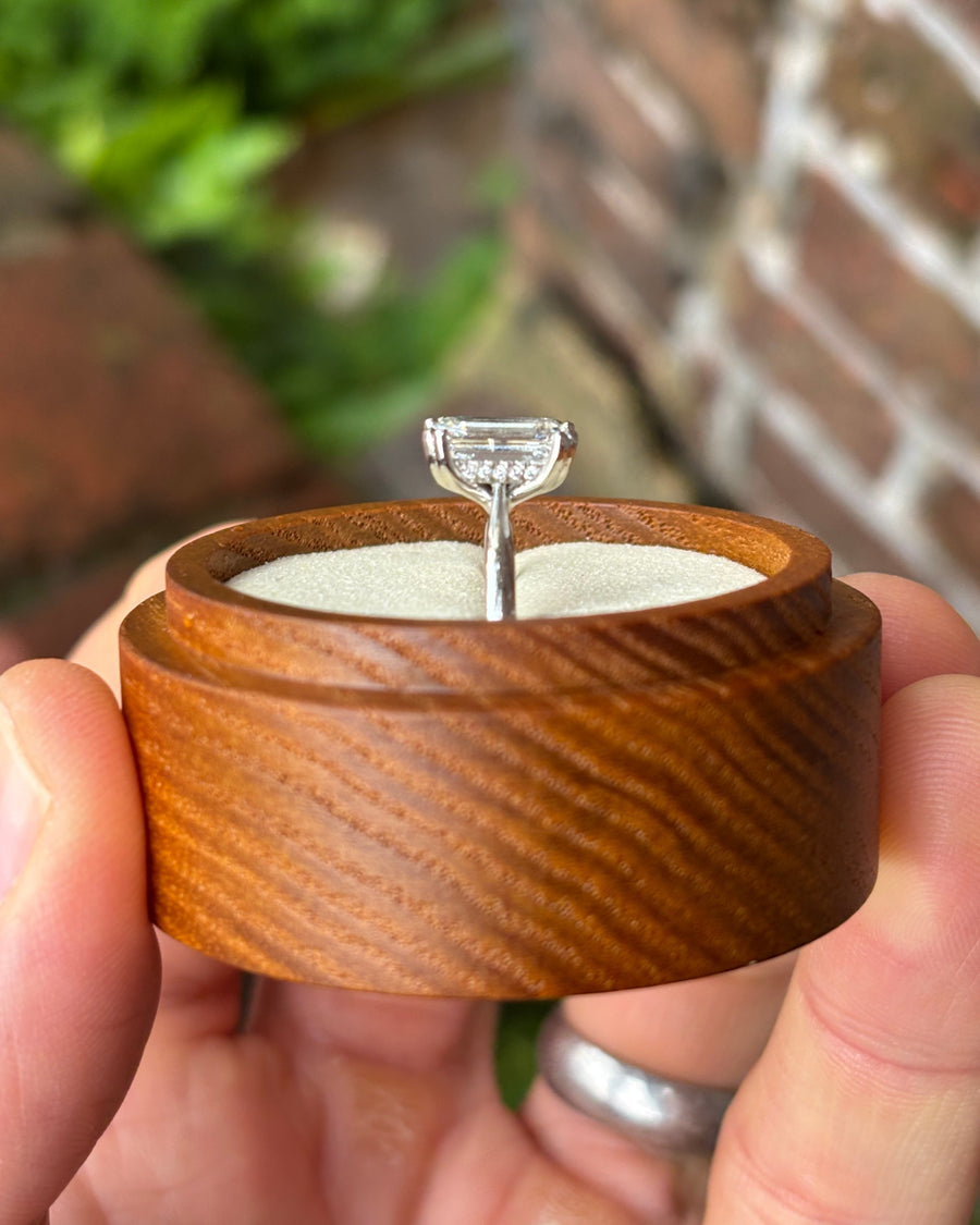 Emerald cut diamond engagement ring with a hidden halo in a white metal setting held in a wooden box.
