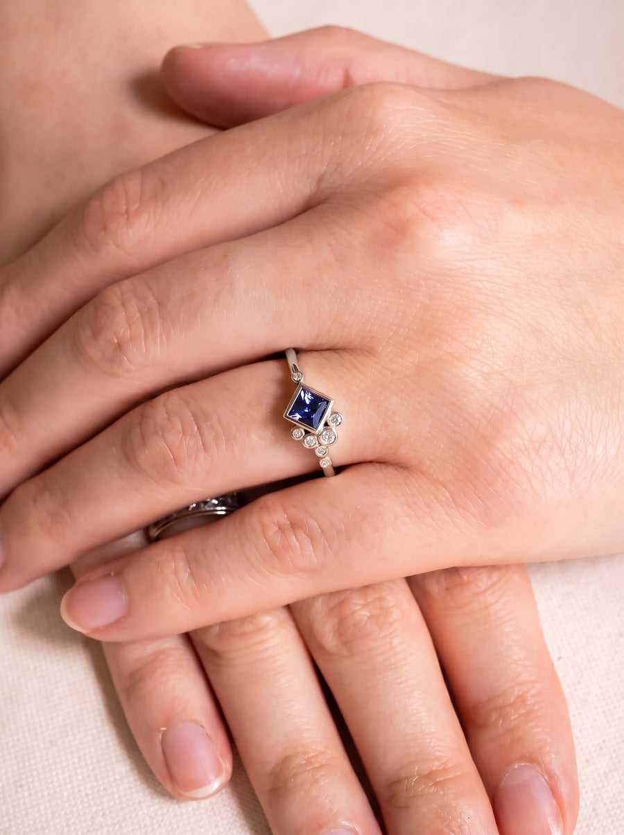Silver engagement ring featuring a square blue gemstone accented by small round diamonds on a thin band.