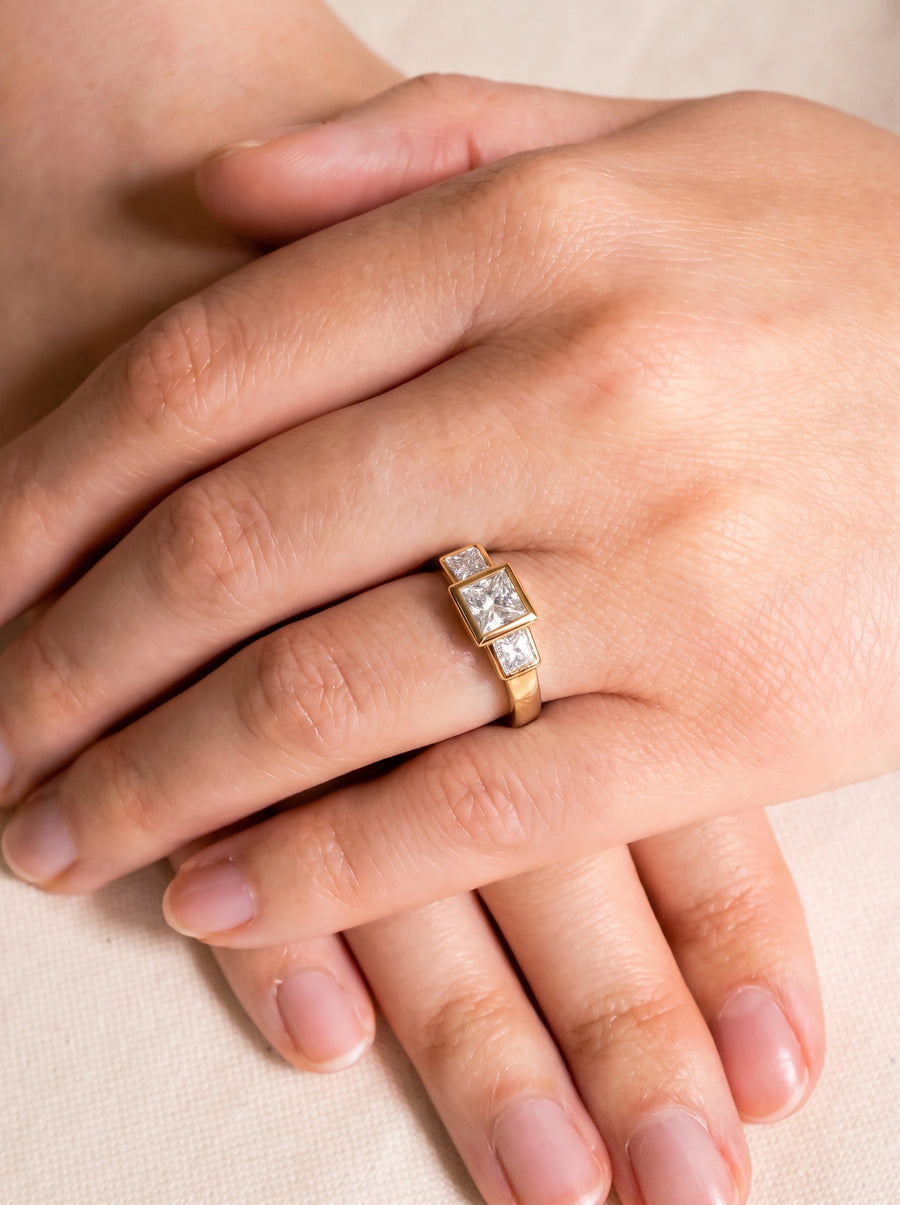 Gold trilogy engagement ring with three princess cut diamonds on a finger.