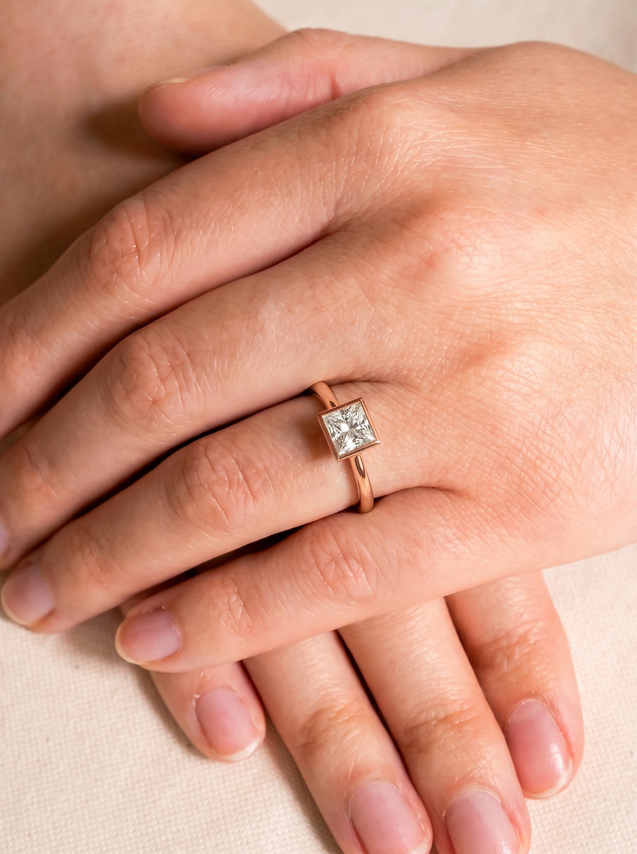 Princess cut diamond engagement ring in a rose gold bezel setting worn on a finger.