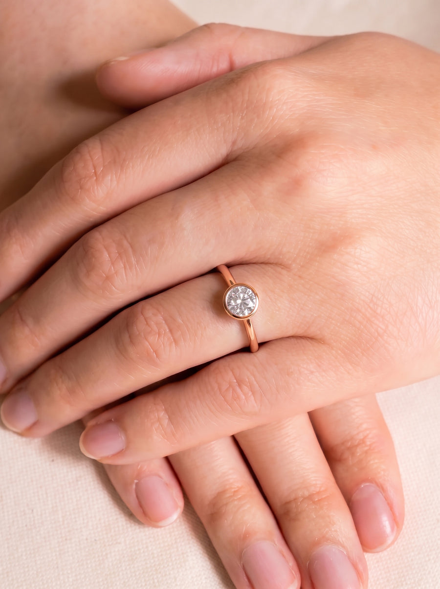 Round cut diamond engagement ring in a rose gold bezel setting worn on a finger.