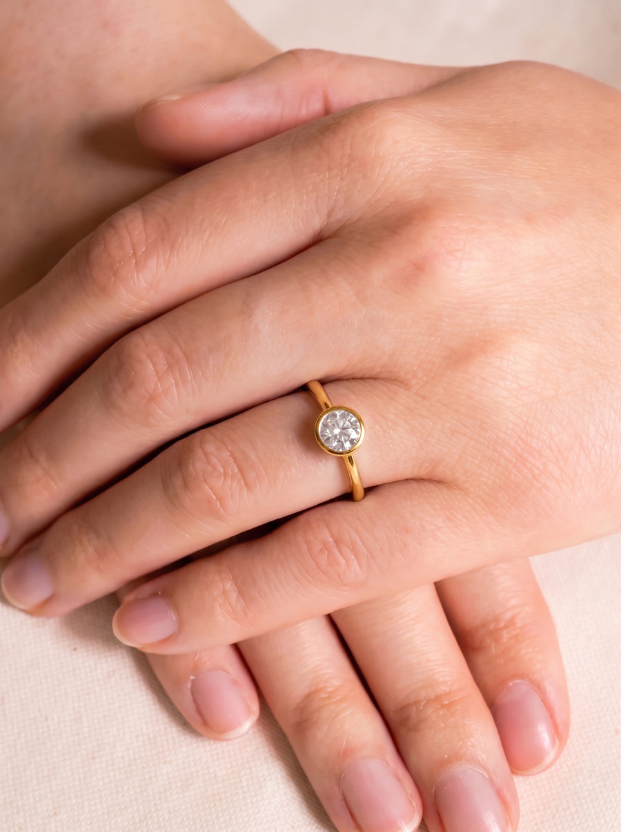 Round cut diamond engagement ring with a gold bezel setting worn on a finger.