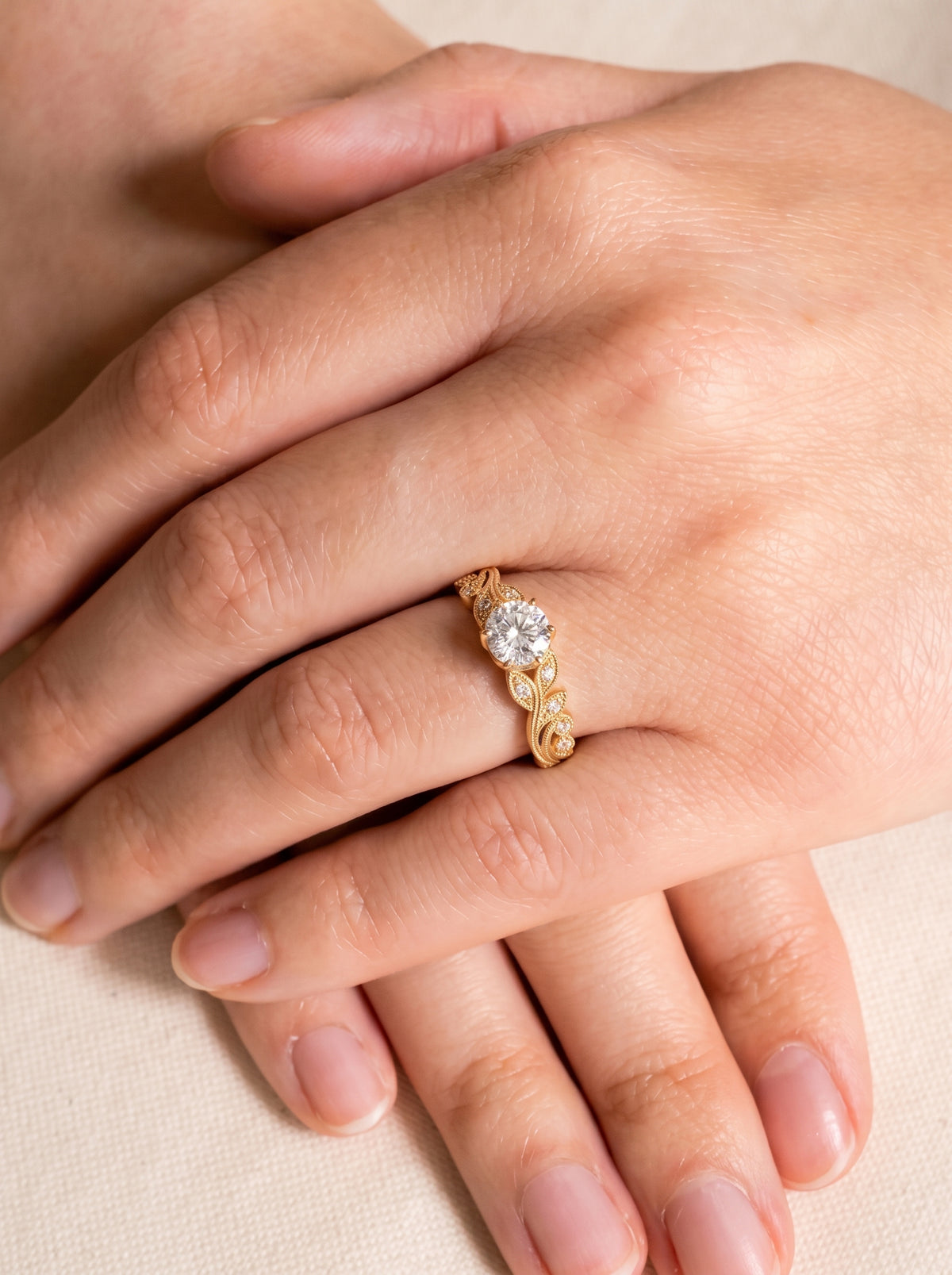 Round cut diamond engagement ring in a yellow gold band with leaf-inspired filigree details worn on a finger.