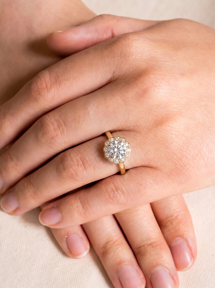 Gold engagement ring with a round diamond cluster, scalloped halo, and heart-shaped side details on a finger.