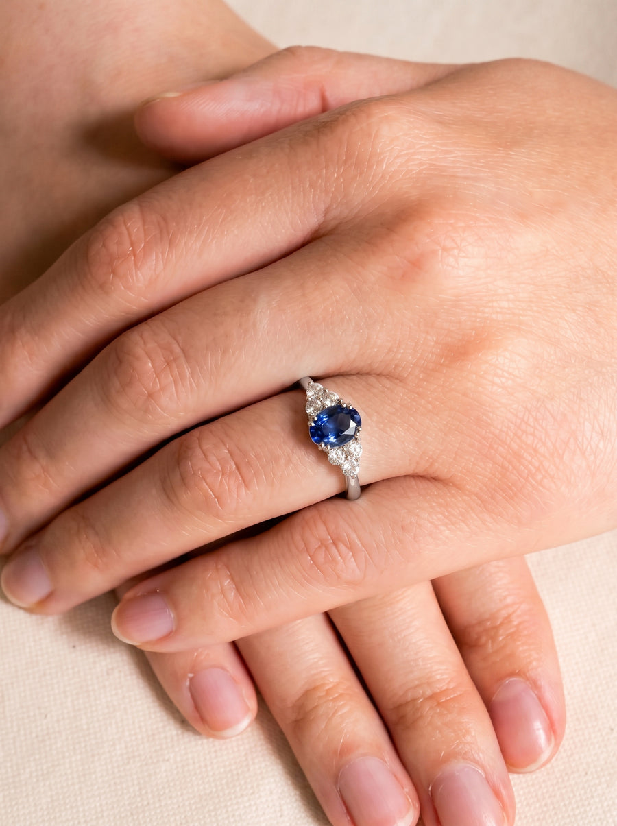 Oval blue sapphire engagement ring with diamond accents on a silver band worn on a finger.