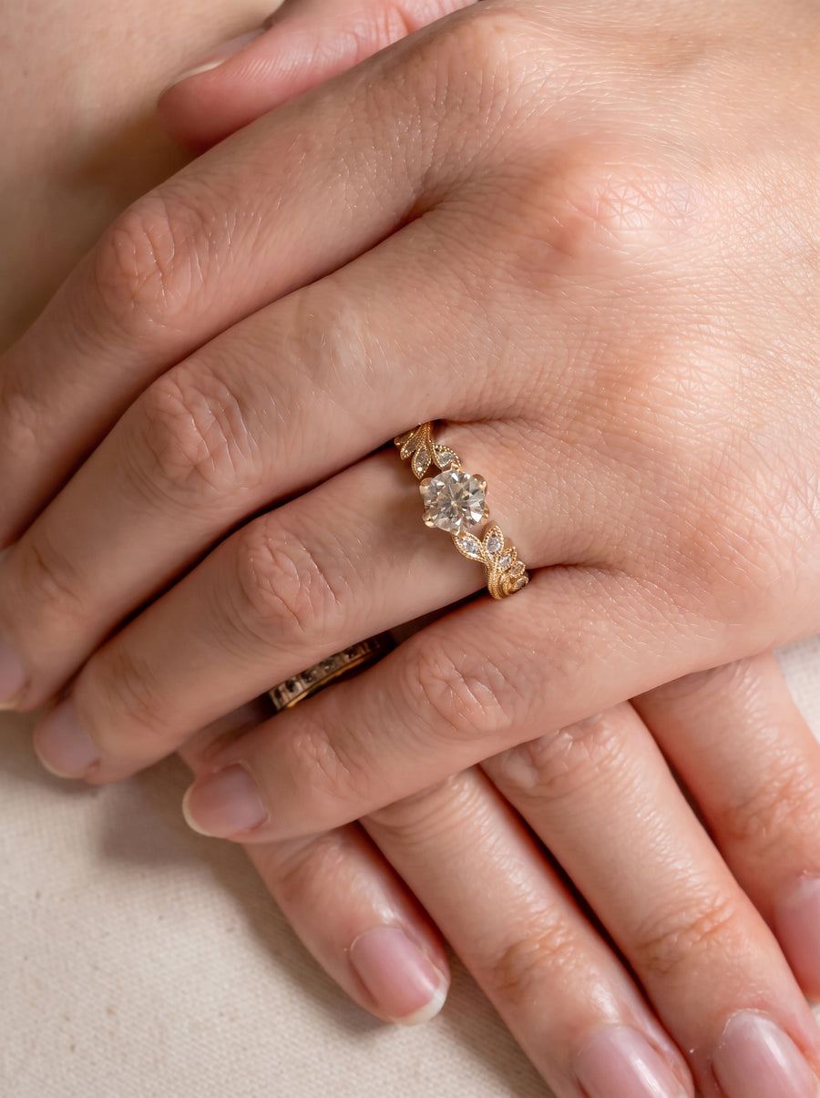 Gold engagement ring with a round diamond, leaf-shaped accents, and small bead details worn on a finger.