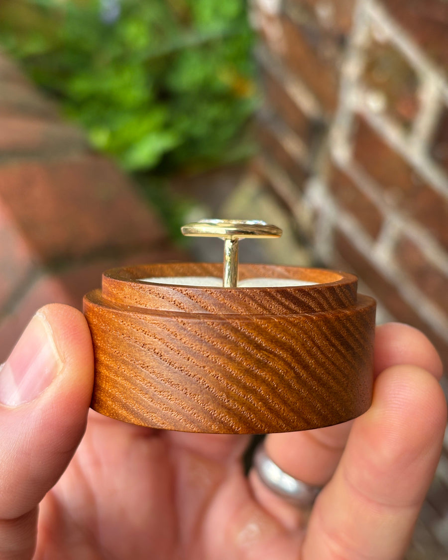 Marquise cut engagement ring with a gold bezel setting held in a round wooden ring box.