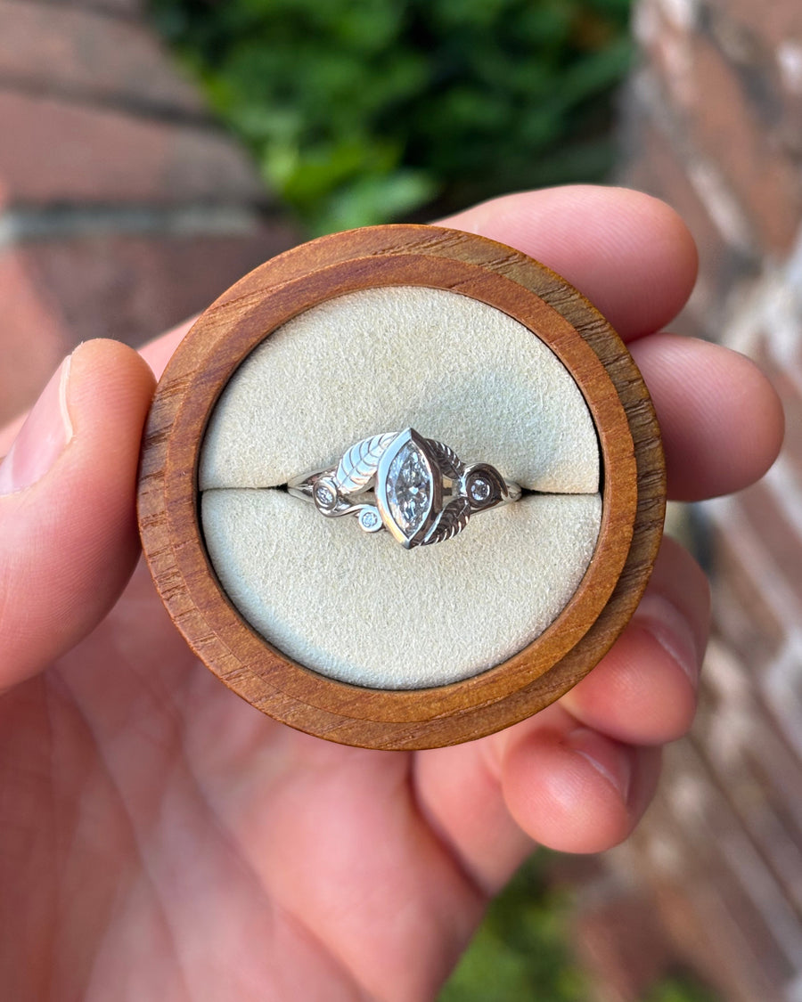 Platinum marquise diamond engagement ring with leaf-inspired metal detailing in a wooden ring box.