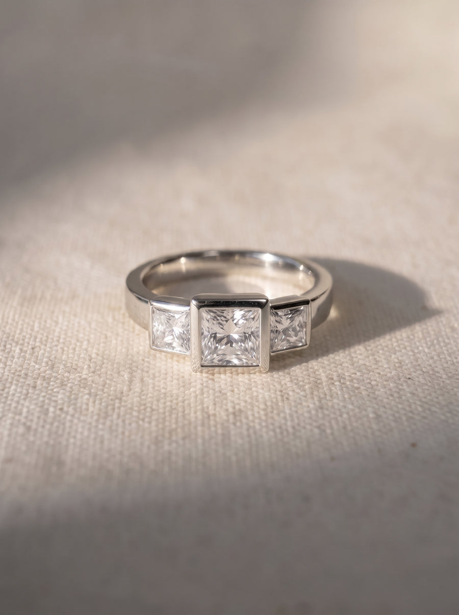 Trilogy engagement ring with three princess cut diamonds in a silver-toned bezel setting on textured fabric.