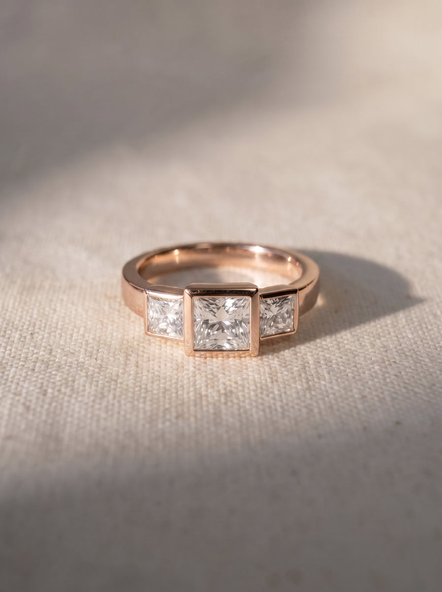 Rose gold trilogy engagement ring featuring three bezel-set princess cut diamonds on a textured fabric background.