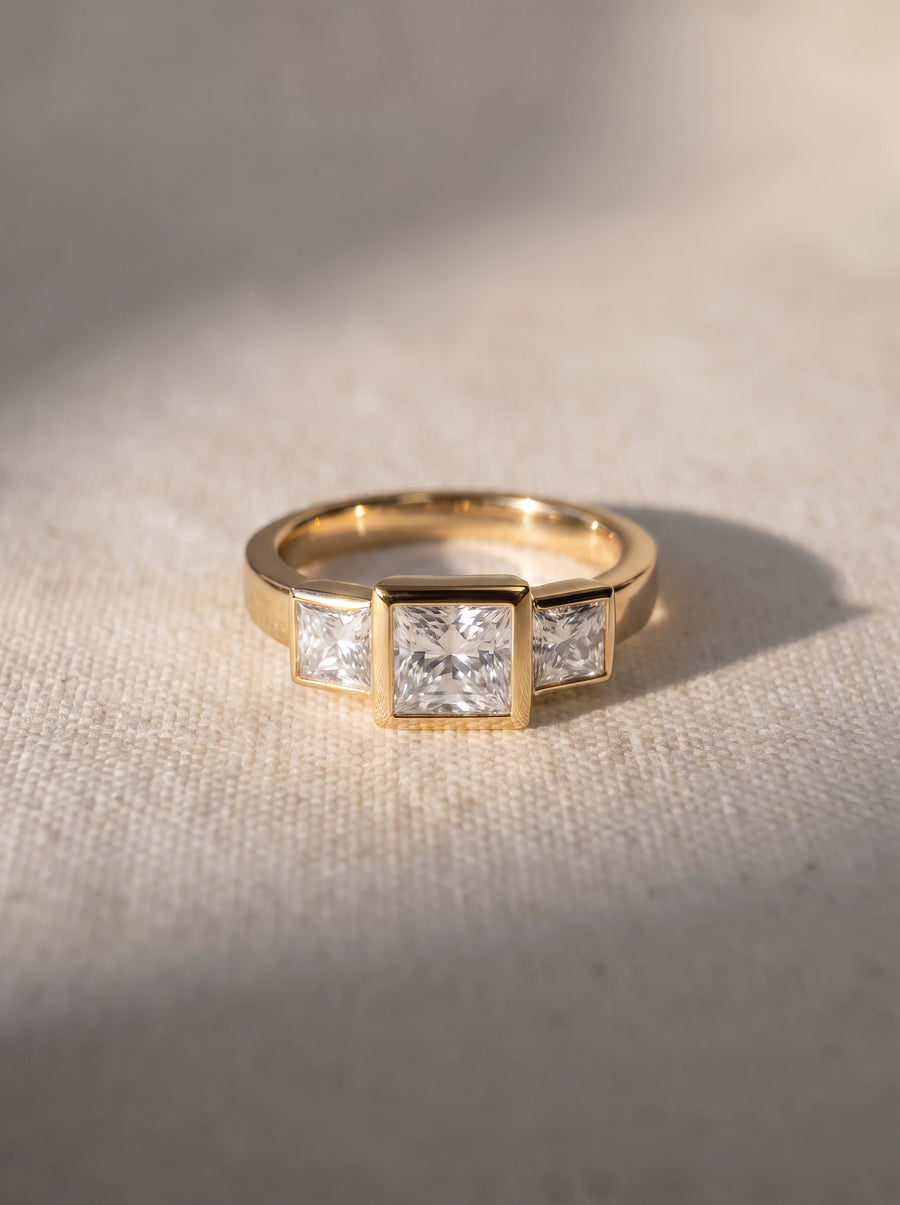 Gold trilogy engagement ring featuring three princess cut diamonds in a bezel setting on a textured surface.