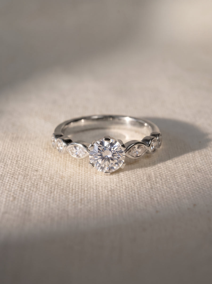Round cut diamond engagement ring in silver metal with marquise and round side stones on a textured fabric.