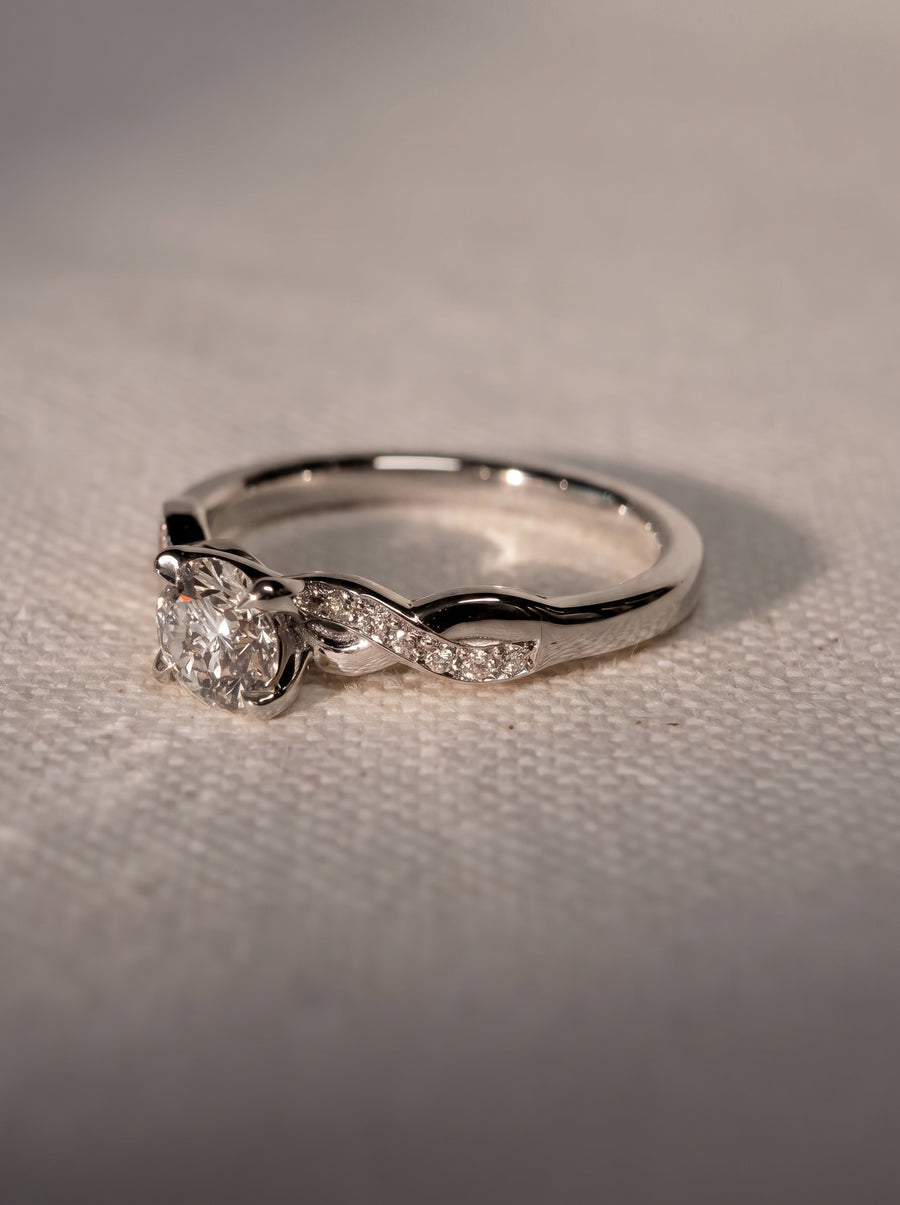 Round cut diamond engagement ring in silver metal with a twisted band featuring small pavé set diamonds.