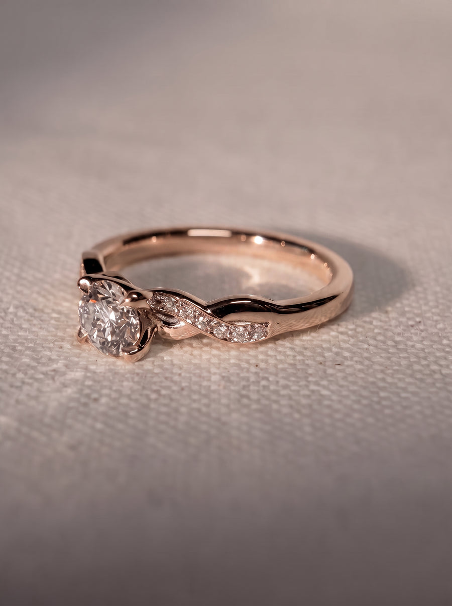 Rose gold engagement ring with a round cut center diamond and a twisted band featuring small accent diamonds.