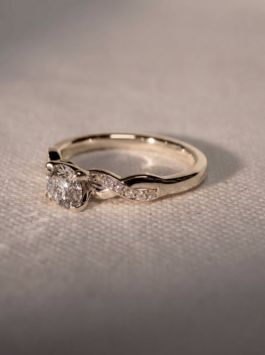 Round cut diamond engagement ring in silver metal with a twisted band featuring small pavé set stones.