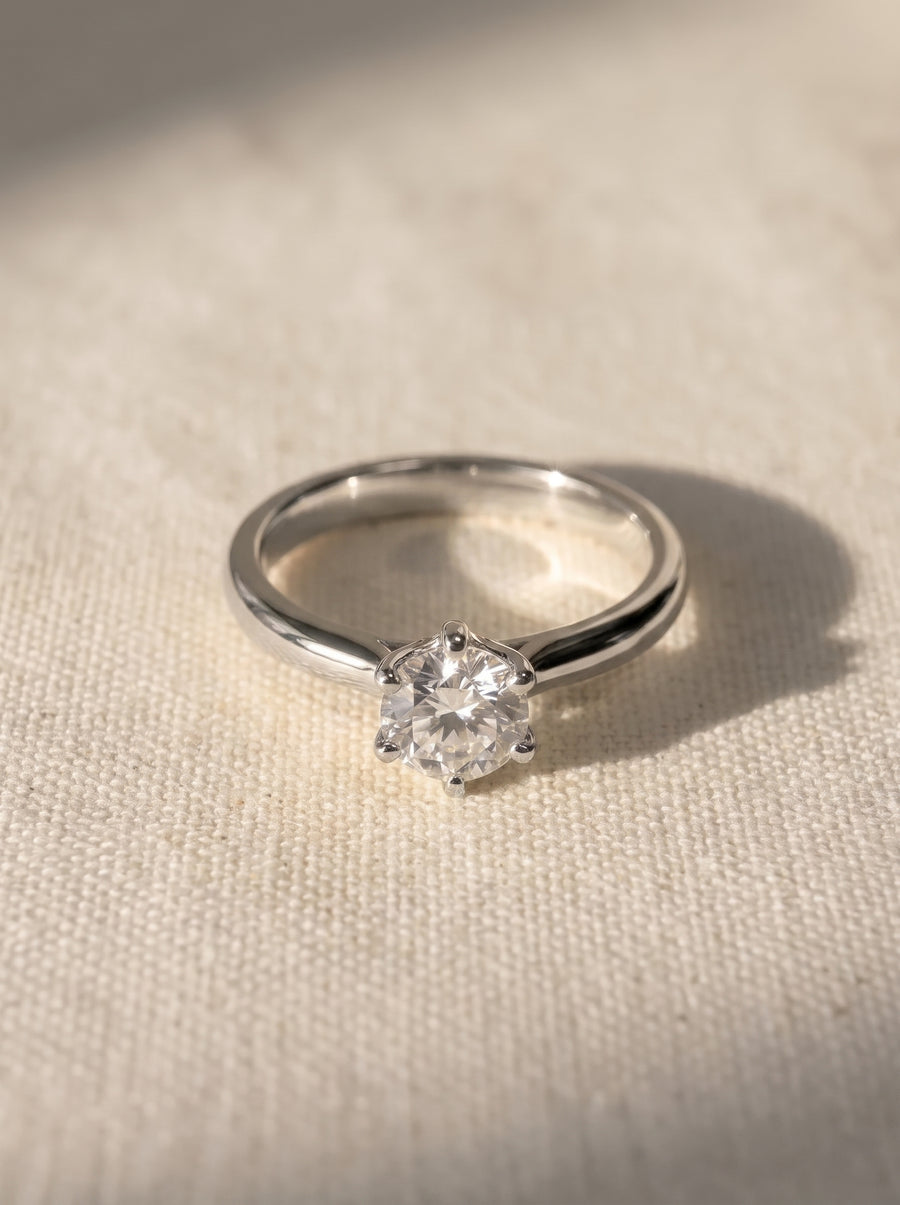 Solitaire engagement ring with a round diamond in a six-claw platinum setting on a textured fabric background.