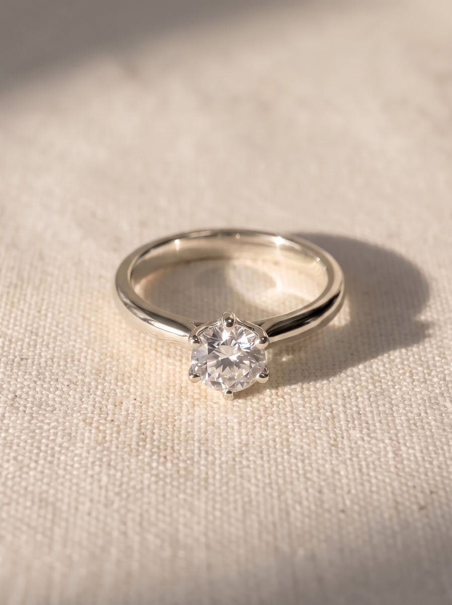 Round diamond solitaire engagement ring in a silver-toned metal setting with six prongs on a textured surface.