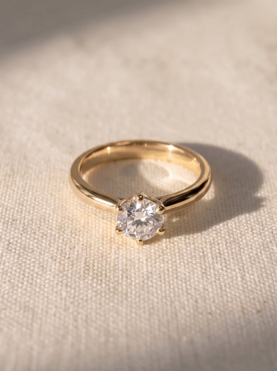 Gold solitaire engagement ring with a round brilliant diamond in a six-claw V setting on textured fabric.