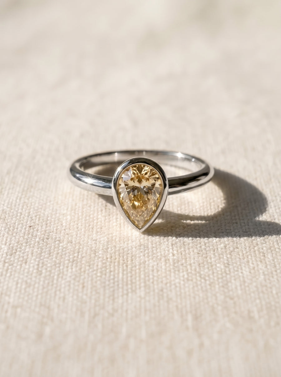 Pear cut yellow diamond solitaire ring in a silver-toned bezel setting on a textured fabric background.