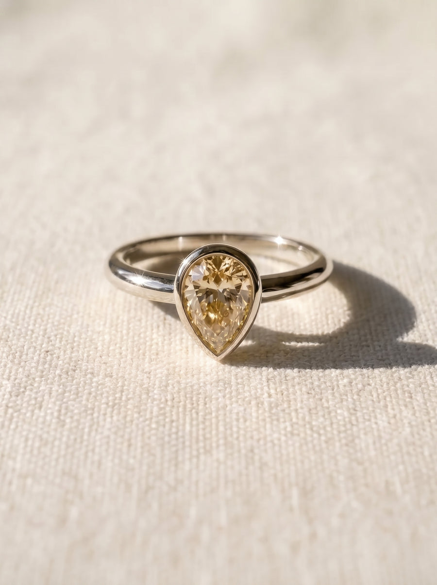 Pear cut yellow diamond solitaire ring in a silver-toned bezel setting on a textured fabric background.