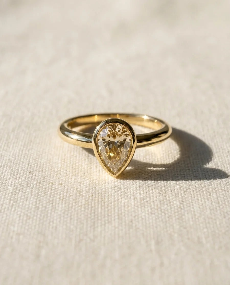 Pear cut diamond solitaire ring in a gold bezel setting on a textured fabric background.