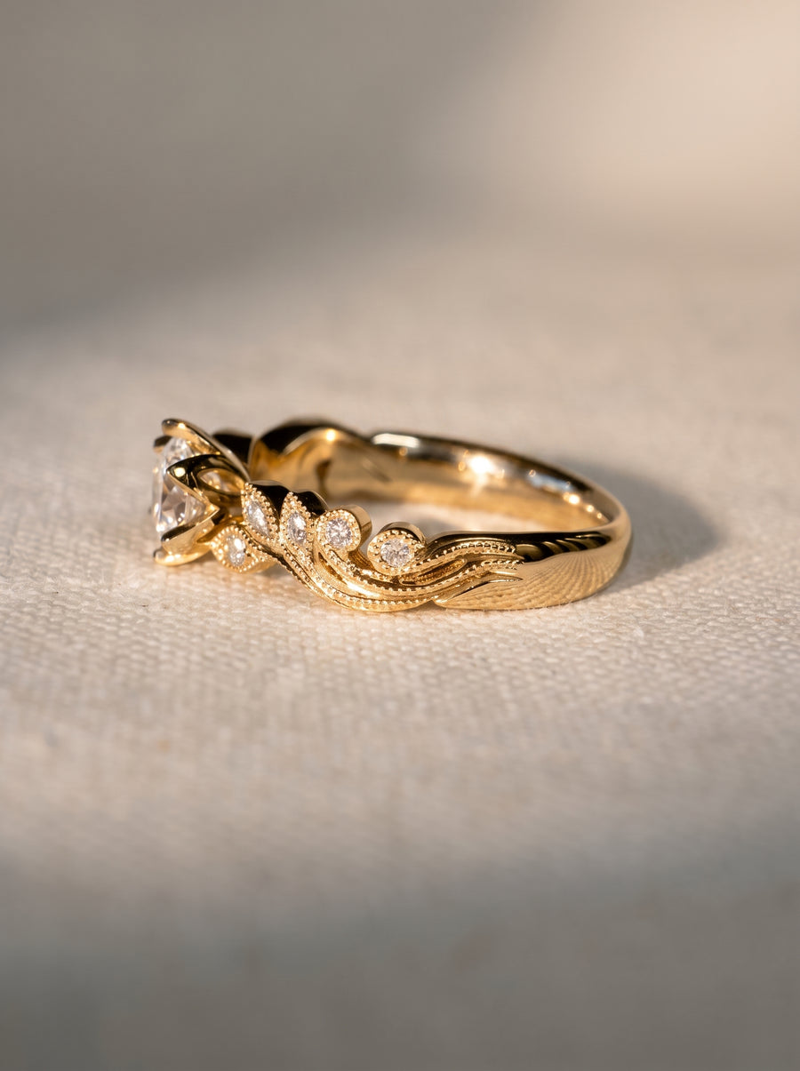 Gold engagement ring with a round diamond, featuring beaded milgrain details and small accent stones on the band.
