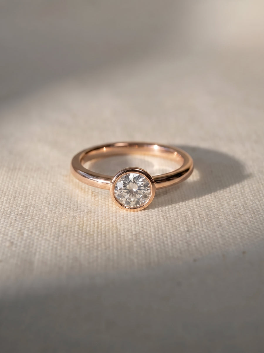 Round cut diamond engagement ring in a rose gold bezel setting on a textured fabric background.