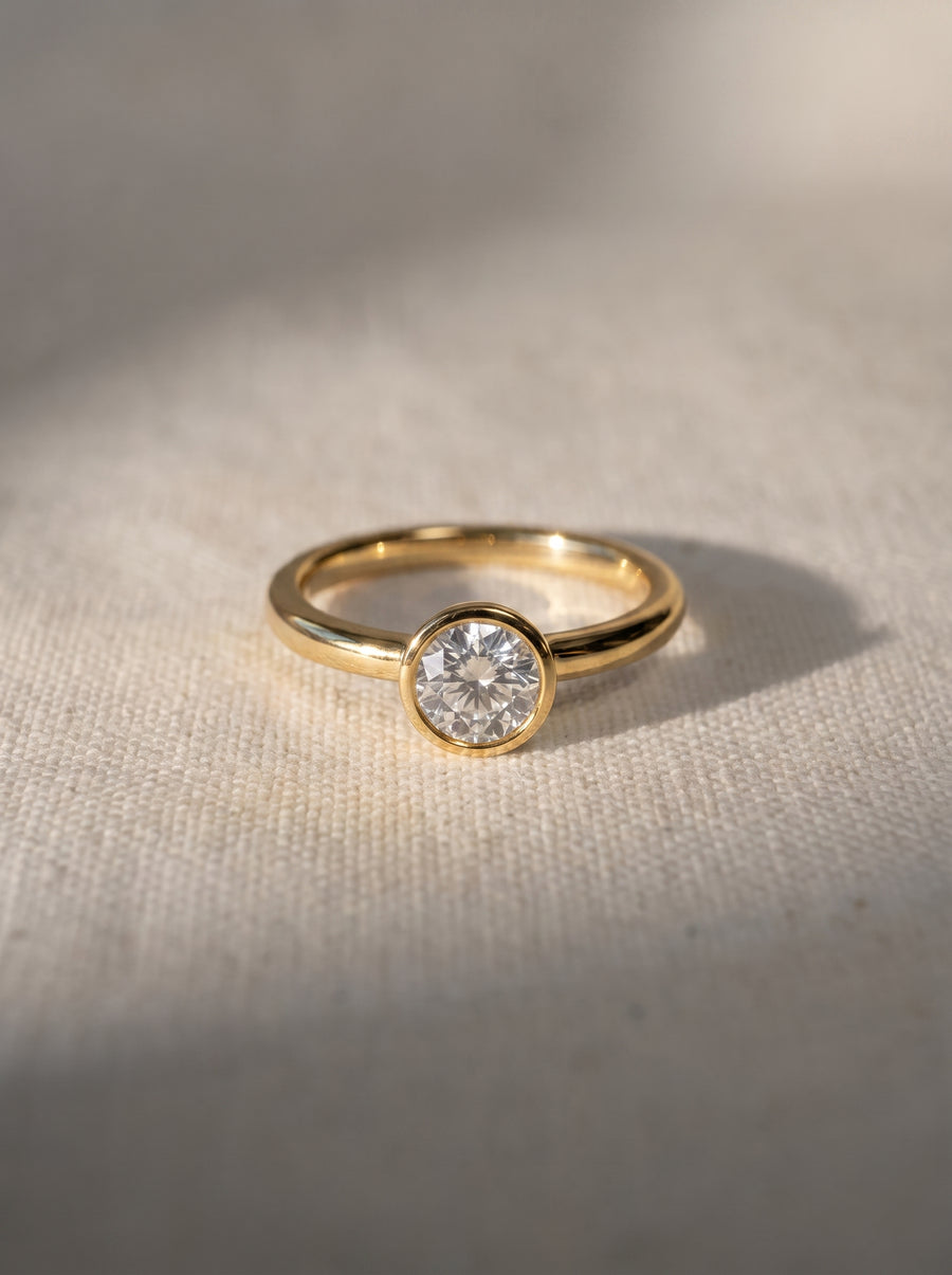 Round cut diamond engagement ring in a gold bezel setting on a textured fabric background.