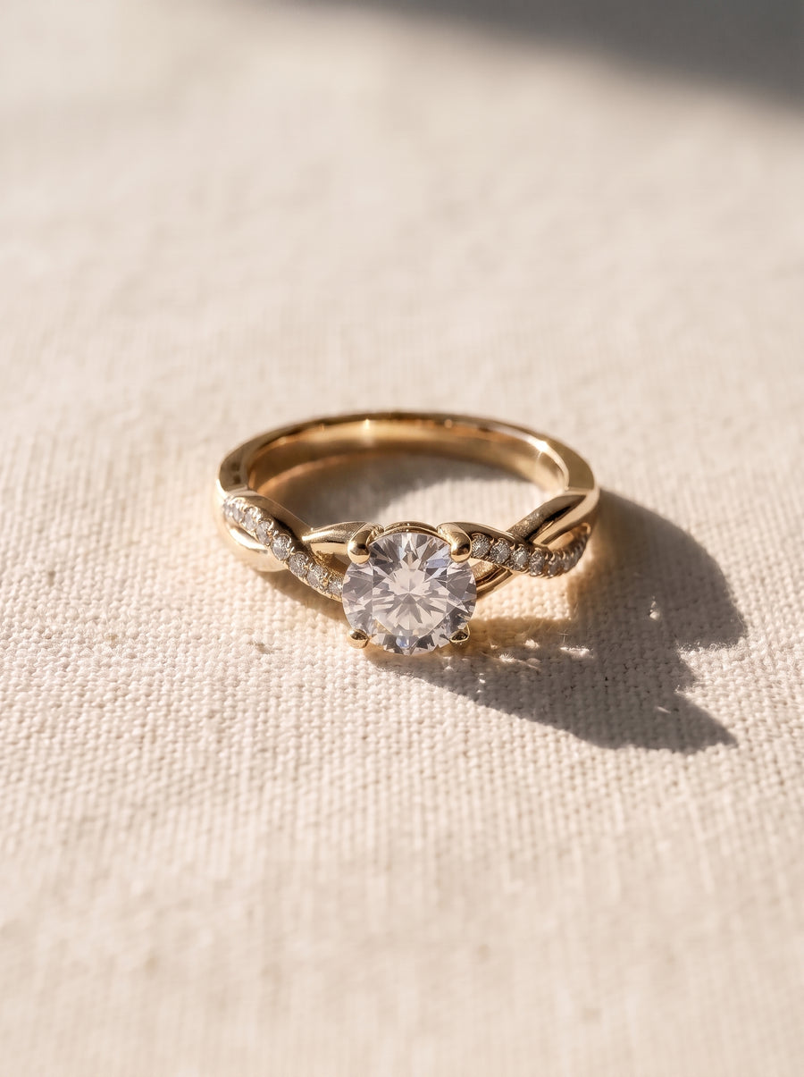 Round cut diamond engagement ring with a gold twist band and pavé accents on a textured fabric surface.
