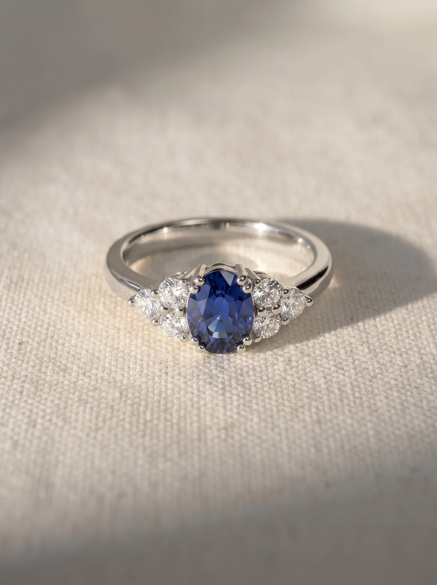 Oval blue sapphire and diamond engagement ring set in a polished silver-tone band on a textured fabric surface.