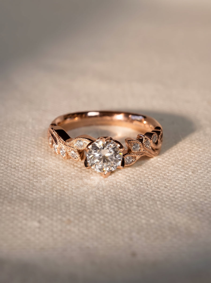Rose gold engagement ring with a round diamond center and leaf-shaped beaded accents on a textured surface.
