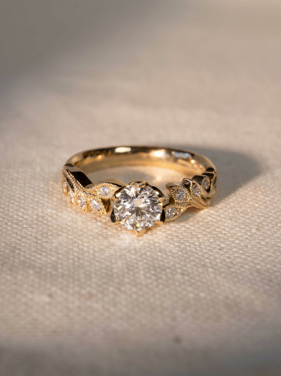 Gold engagement ring with a round diamond center stone and leaf-inspired beaded diamond accents on the band.