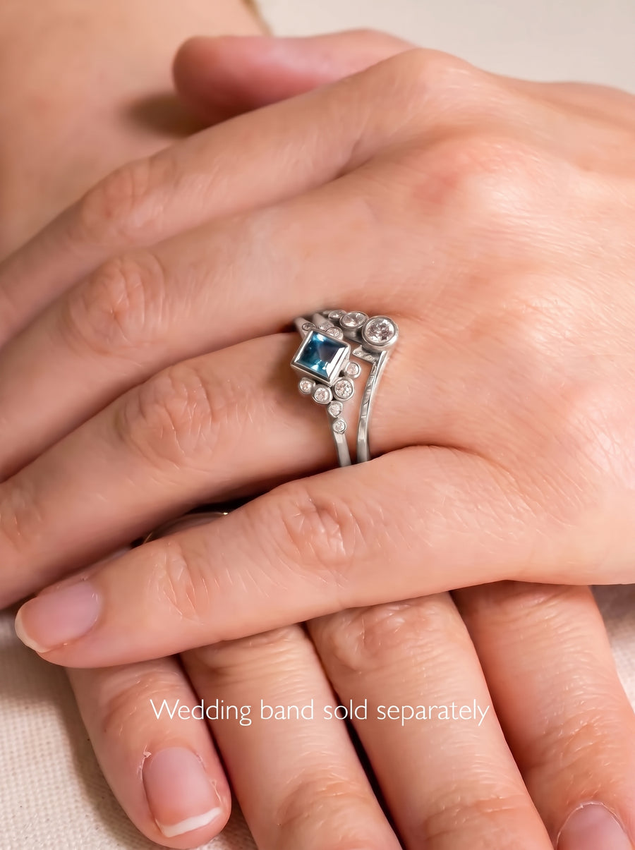 Silver engagement ring with a square blue gemstone and small round diamonds on a finger.