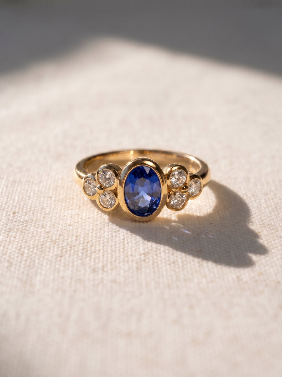 Oval blue sapphire engagement ring in yellow gold with trefoil diamond clusters on a textured fabric background.
