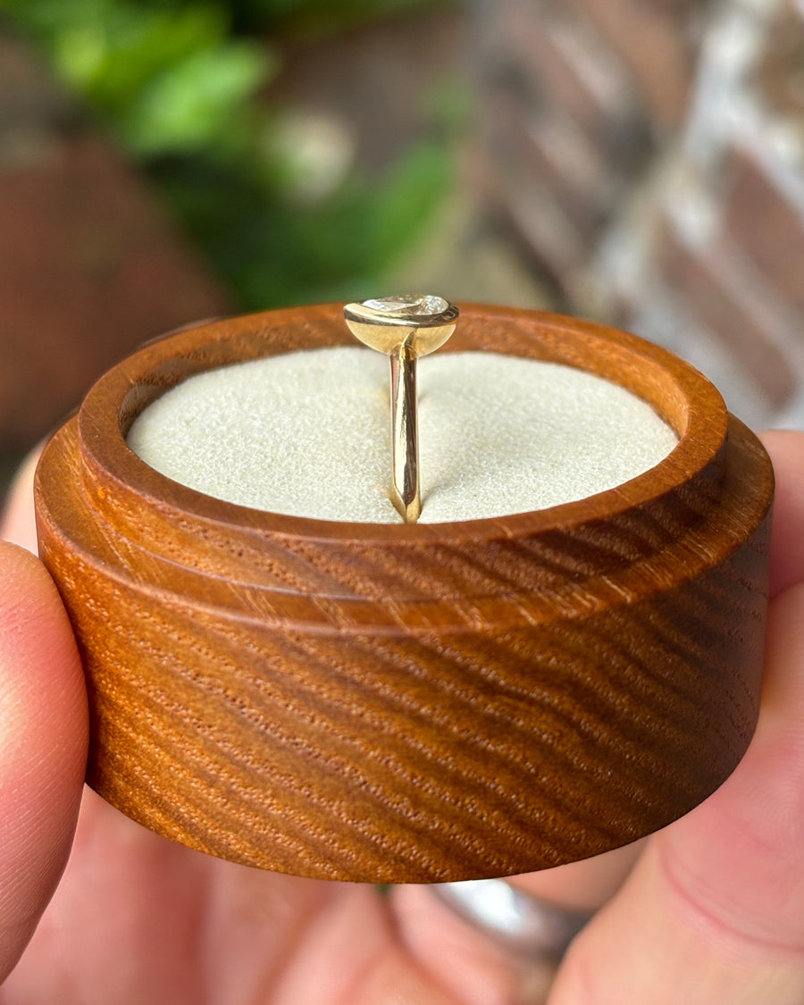 Pear cut diamond engagement ring in a gold bezel setting held in a round wooden box.