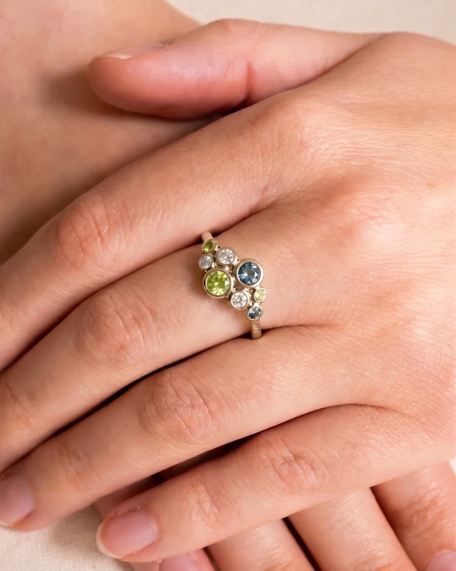 Gold cluster ring featuring bezel-set green and teal sapphires and clear diamonds on a finger.