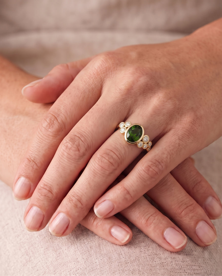 Gold dress ring featuring an oval green gemstone surrounded by small round diamonds on a finger.