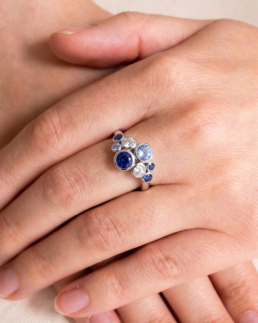 Platinum cluster ring featuring round blue sapphires and diamonds in a bubble-style bezel setting on a finger.