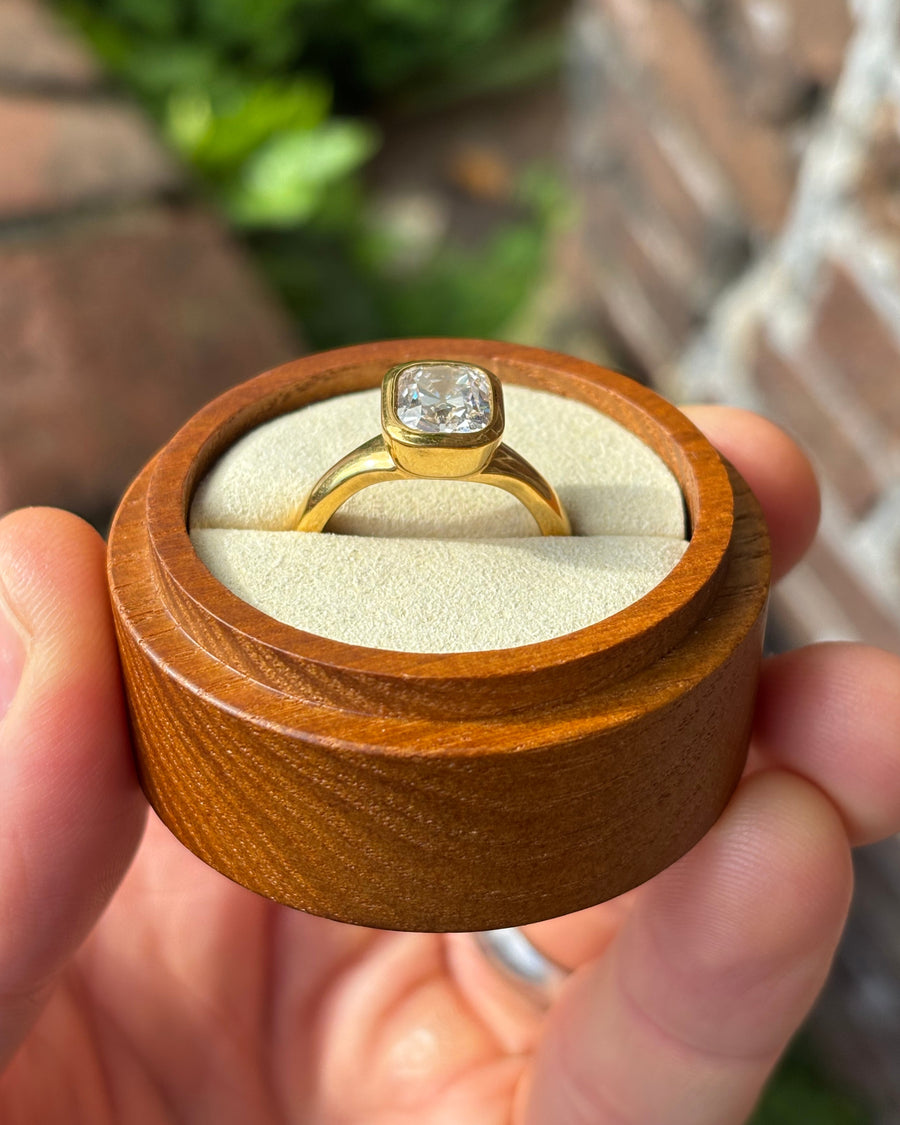 Gold engagement ring with a cushion cut diamond in a bezel setting held in a wooden box.