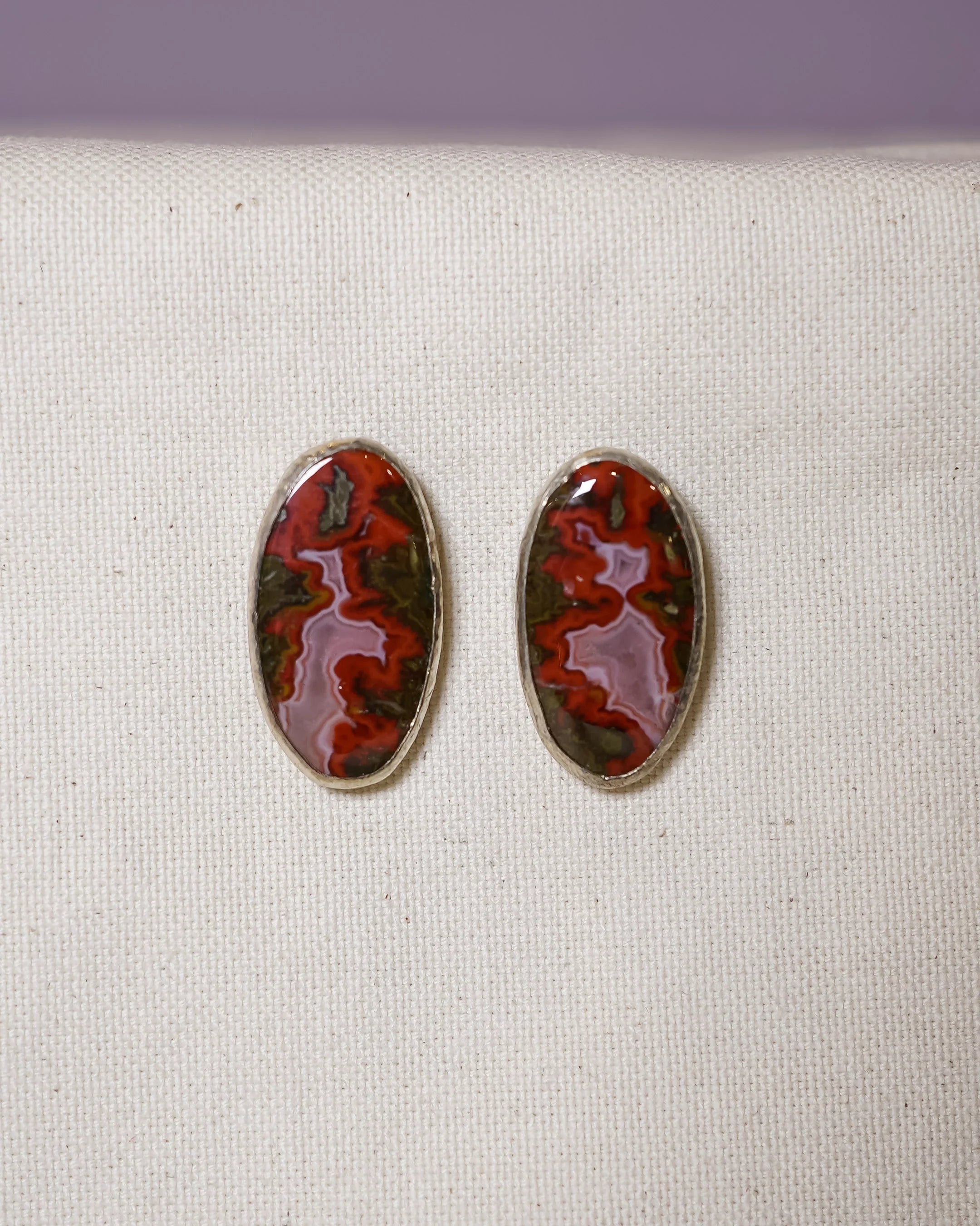 Oval gemstone stud earrings with red and brown organic patterns on cream fabric background