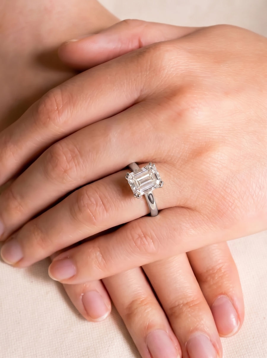 Emerald-cut diamond solitaire engagement ring with a cathedral setting on a silver-toned band worn on a finger.