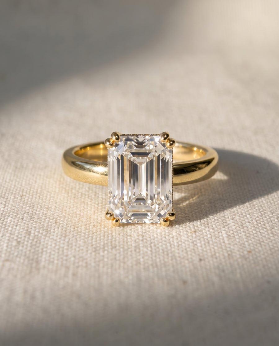 Emerald-cut solitaire diamond engagement ring set in a polished yellow gold cathedral band.