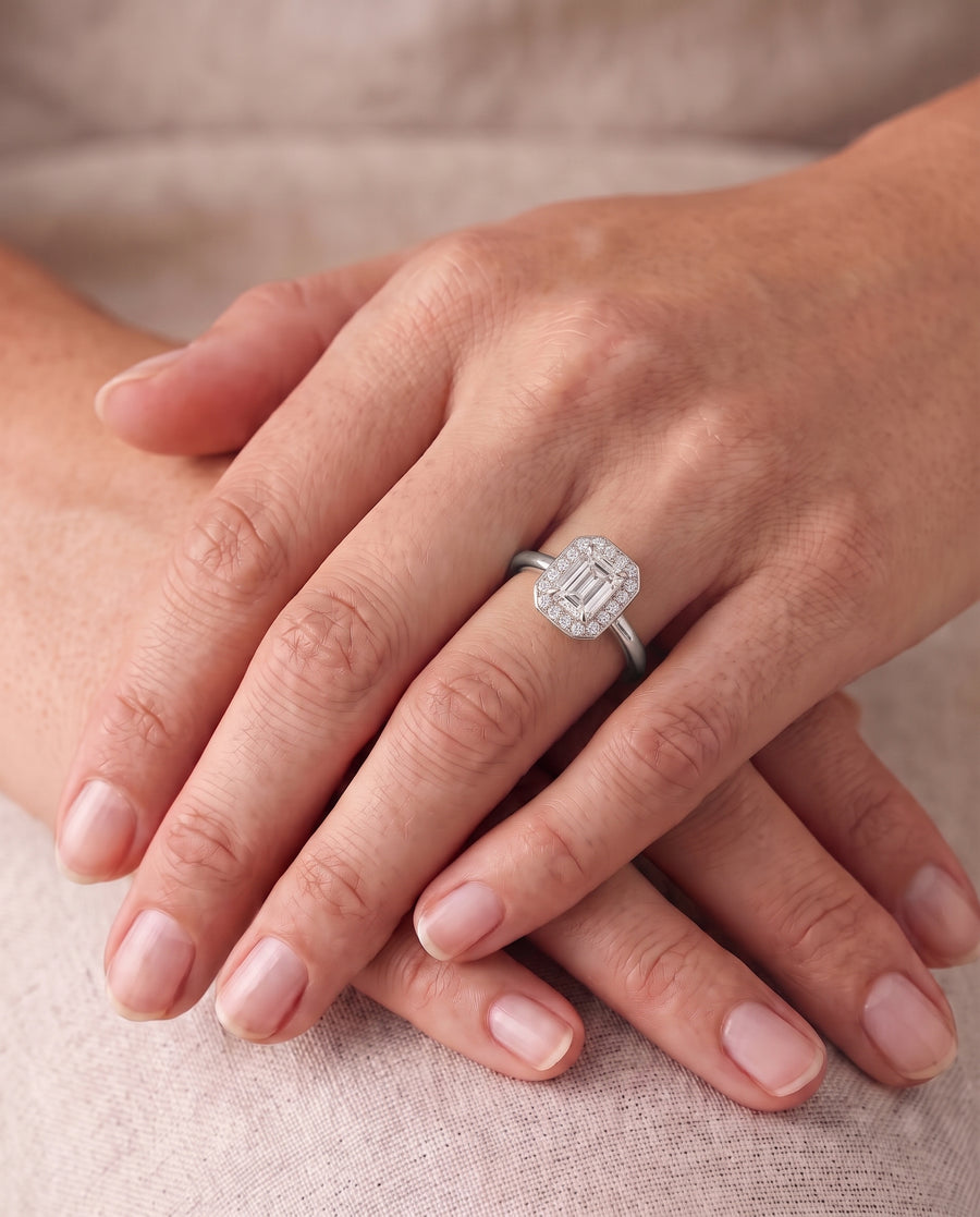 Emerald-cut diamond engagement ring with a diamond halo on a silver band worn on a finger.