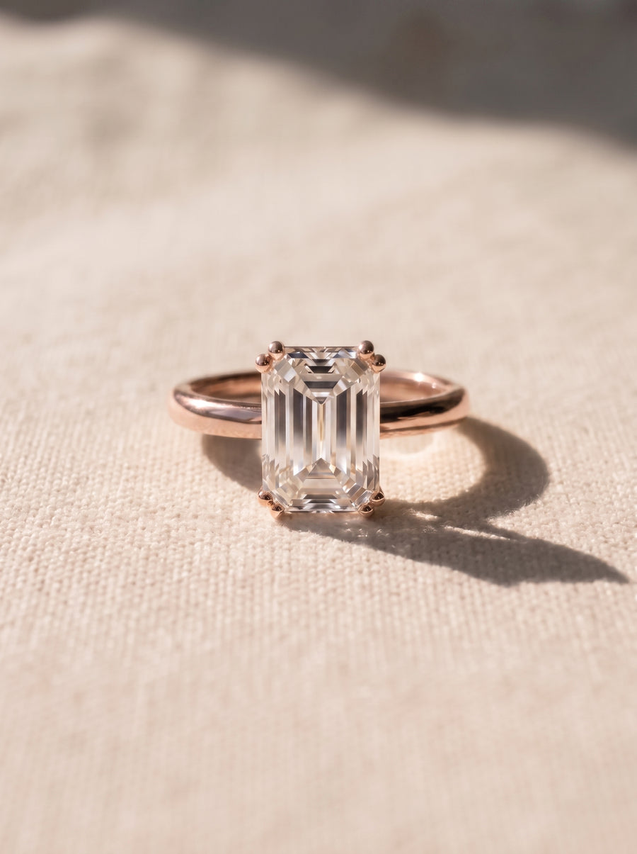 Emerald cut diamond engagement ring set in a polished rose gold band on a textured fabric background.
