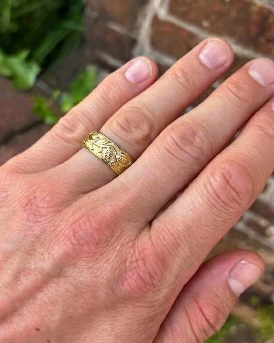 Gold wedding band featuring an engraved leaf motif worn on a finger.