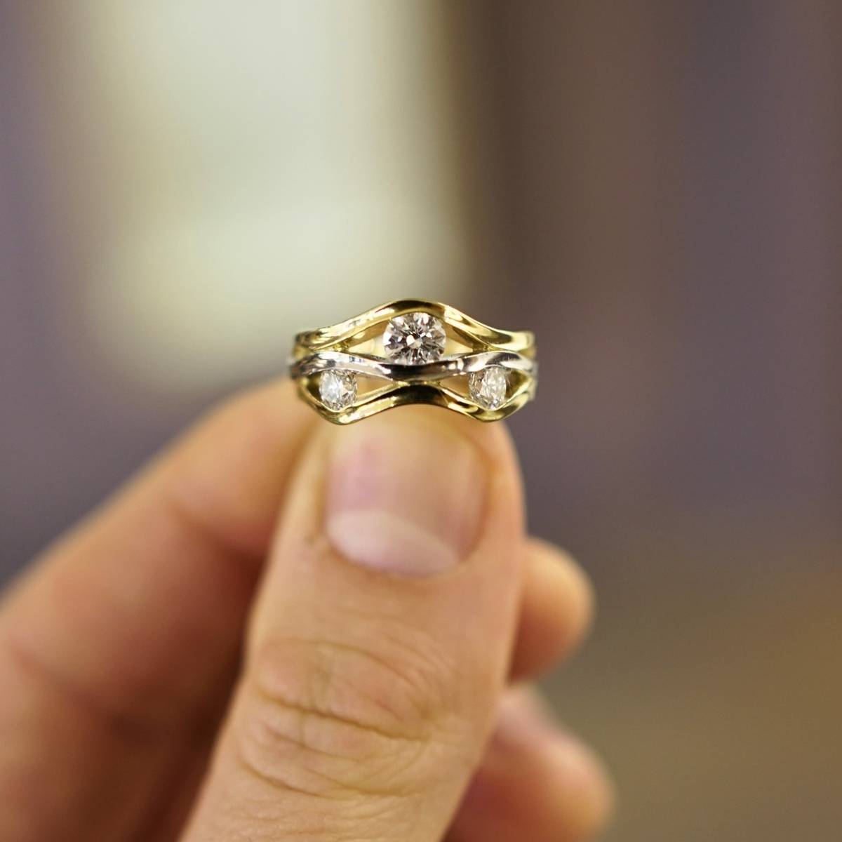 A gold ring with three round diamonds, held between fingers against a softly blurred background.